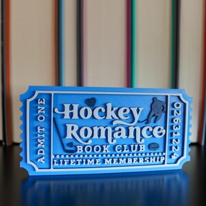 May include: A blue and white 3D-printed ticket with the words "Hockey Romance Book Club" and "Lifetime Membership." The ticket has a hockey player silhouette and a heart symbol. The ticket reads "Admit One" and has a serial number.
