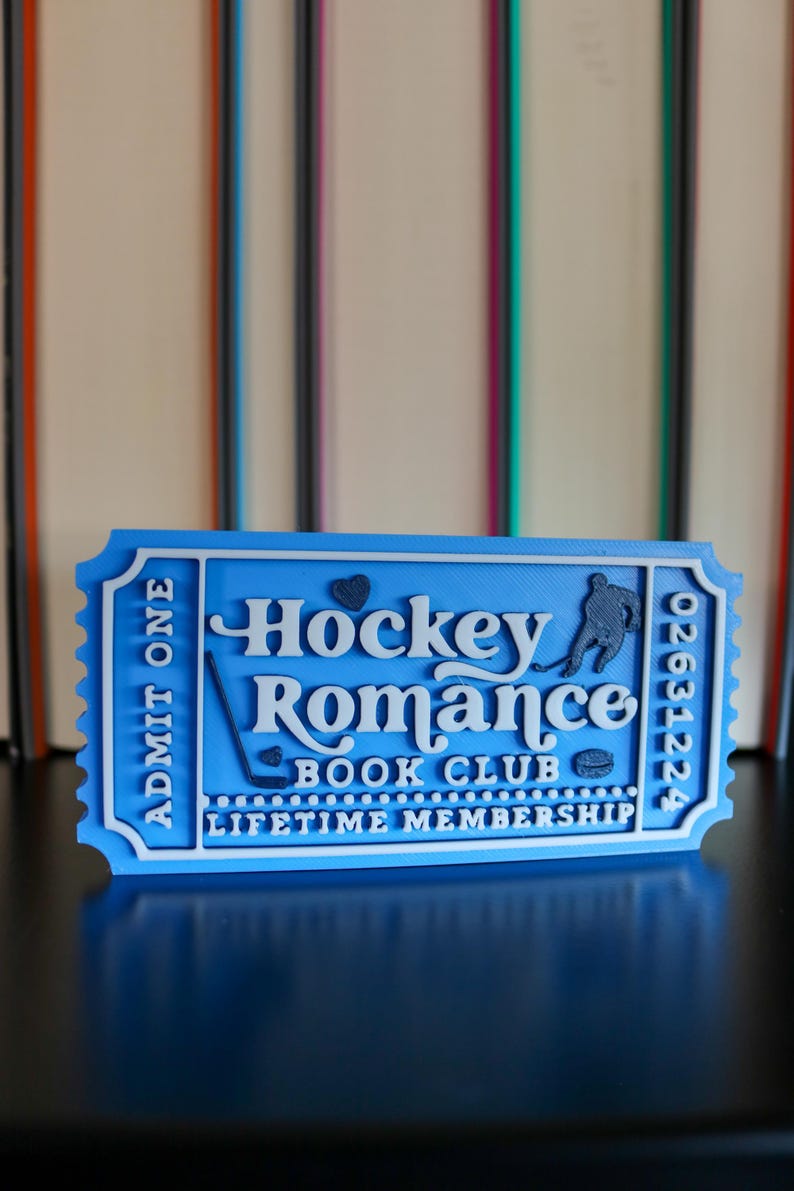 May include: A blue and white 3D-printed ticket with the words "Hockey Romance Book Club" and "Lifetime Membership." The ticket has a hockey player silhouette and a heart symbol. The ticket reads "Admit One" and has a serial number.