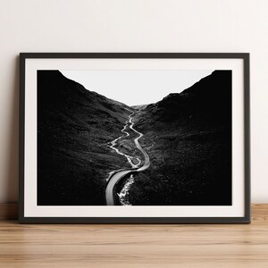 May include: A black and white photograph of a winding road through a mountain pass. The road is surrounded by hills and the sky is visible in the distance.