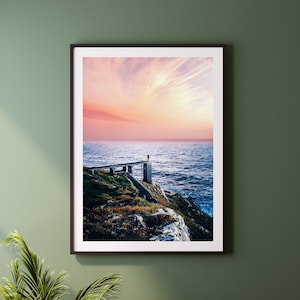 Puede incluir: Una fotografía de una costa rocosa con una persona de pie en una estructura de hormigón que domina el océano. El cielo es de un rosa y naranja vibrantes, con nubes que reflejan la puesta de sol.