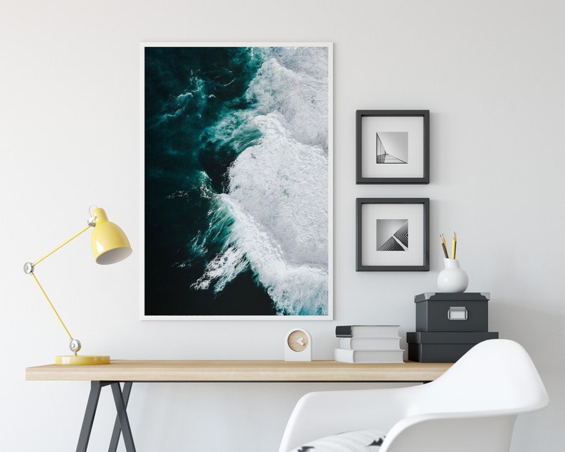 May include: A framed art print of ocean waves in shades of teal and white. The artwork is displayed on a white wall above a wooden desk with a yellow desk lamp. Two framed black and white prints and a small clock are also on the wall.