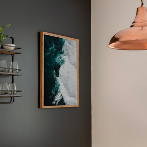 May include: A framed ocean wave art print hangs on a dark gray wall, next to a metal and wood shelf with glasses and decorative bowls. A copper pendant light hangs from the ceiling, illuminating the space. The artwork features shades of teal and white.
