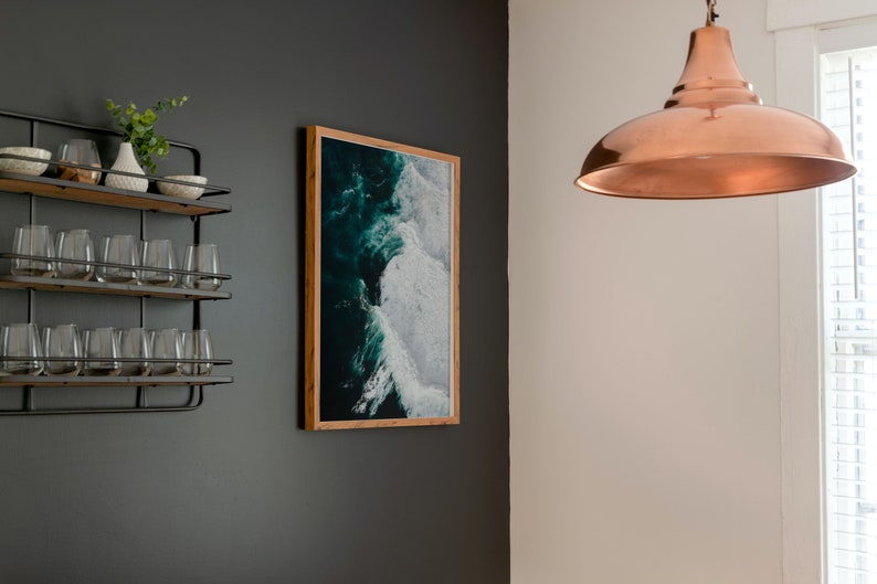 May include: A framed ocean wave art print hangs on a dark gray wall, next to a metal and wood shelf with glasses and decorative bowls. A copper pendant light hangs from the ceiling, illuminating the space. The artwork features shades of teal and white.
