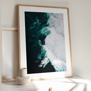 May include: A framed art print featuring an aerial view of ocean waves. The artwork showcases dark teal water contrasting with white foamy crests. The print is set in a light wood frame, leaning against a white wall.