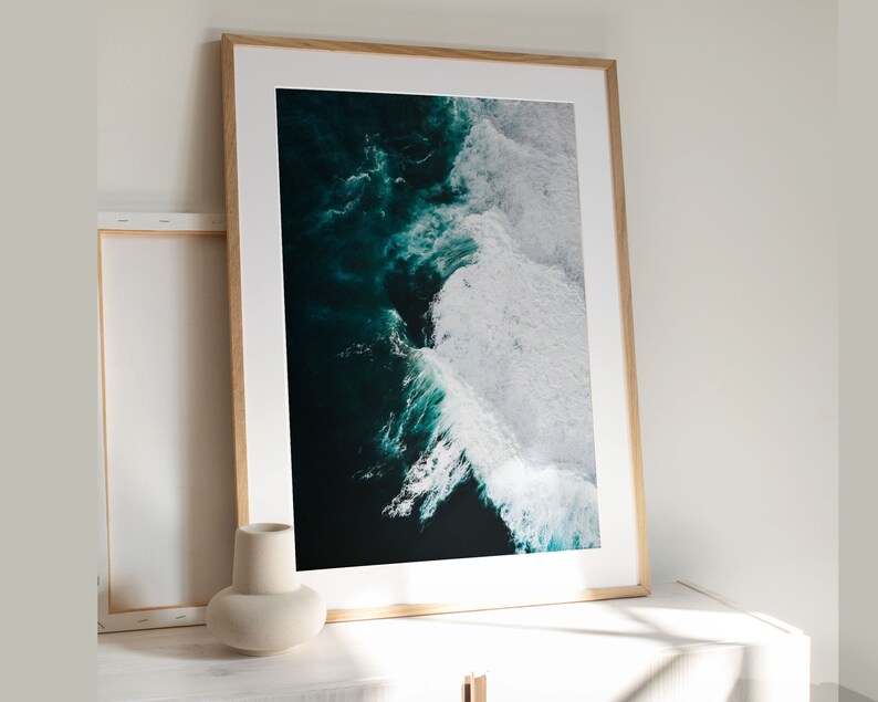 May include: A framed art print featuring an aerial view of ocean waves. The artwork showcases dark teal water contrasting with white foamy crests. The print is set in a light wood frame, leaning against a white wall.
