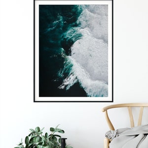 May include: A framed art print featuring an aerial view of ocean waves. The artwork showcases dark teal water transitioning to white foamy crests. The print is in a black frame, displayed against a white wall, with a woven basket and a black vase in the foreground.