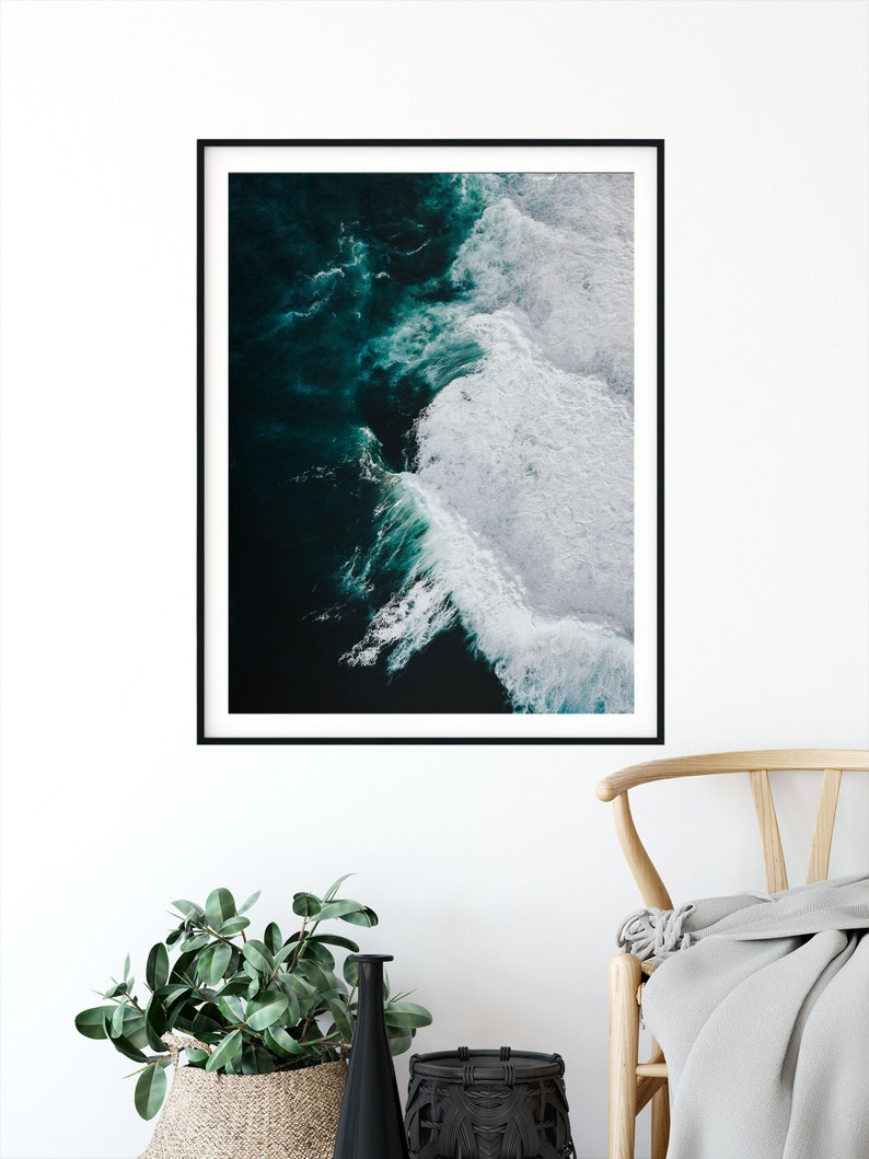 May include: A framed art print featuring an aerial view of ocean waves. The artwork showcases dark teal water transitioning to white foamy crests. The print is in a black frame, displayed against a white wall, with a woven basket and a black vase in the foreground.