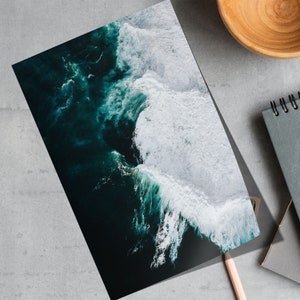 May include: A print featuring an aerial view of ocean waves. The image showcases dark teal water with white foamy crests. A wooden bowl, a spiral-bound notebook, and a pencil are also visible.