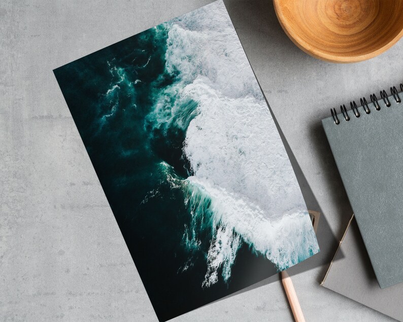 May include: A print featuring an aerial view of ocean waves. The image showcases dark teal water with white foamy crests. A wooden bowl, a spiral-bound notebook, and a pencil are also visible.