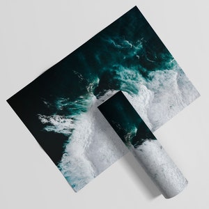 May include: Two prints featuring an aerial view of ocean waves. One print is flat, while the other is rolled up. The ocean is a deep teal color, with white foamy waves crashing against the shore. The prints are on a white background.