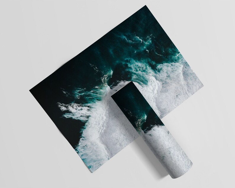 May include: Two prints featuring an aerial view of ocean waves. One print is flat, while the other is rolled up. The ocean is a deep teal color, with white foamy waves crashing against the shore. The prints are on a white background.