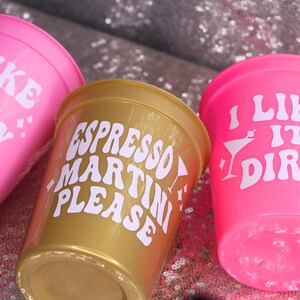 May include: Three plastic cups in pink and gold. The pink cups have the text "I LIKE IT DIRTY" and "I LIKE IT PARTY" in white. The gold cup says "ESPRESSO MARTINI PLEASE" in white. The cups are on a sparkly surface.