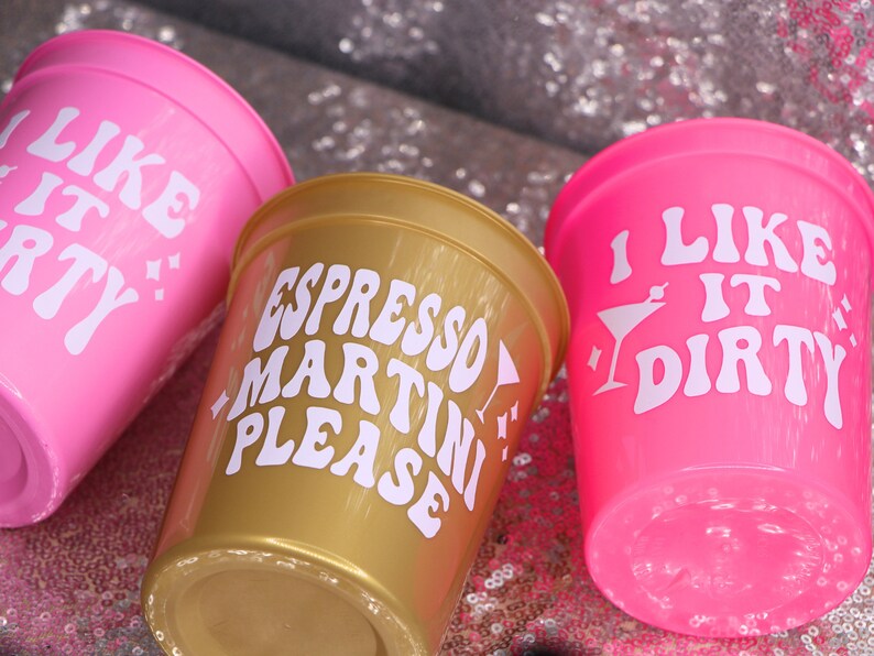 May include: Three plastic cups in pink and gold. The pink cups have the text "I LIKE IT DIRTY" and "I LIKE IT PARTY" in white. The gold cup says "ESPRESSO MARTINI PLEASE" in white. The cups are on a sparkly surface.