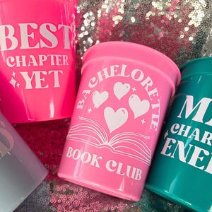 May include: A collection of colorful plastic cups with white text. One cup is bright pink and says "BEST CHAPTER YET". Another pink cup says "BACHELORETTE BOOK CLUB". A teal cup says "MAIN CHARACTER ENERGY".