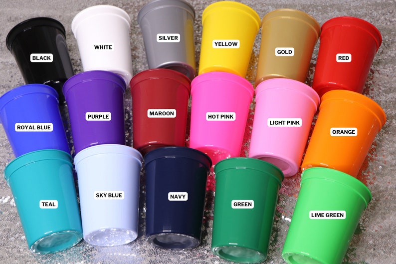 Op de afbeelding: Een verzameling plastic bekers in verschillende kleuren, waaronder zwart, wit, zilver, geel, goud, rood, paars, kastanjebruin, felroze, lichtroze, koningsblauw, oranje, hemelsblauw, marineblauw, blauwgroen, groen en limoengroen. Elke beker is voorzien van een label met de kleurnaam.