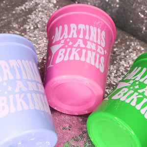 Op de afbeelding: Drie plastic bekers in pastelkleuren: lichtblauw, roze en groen. Elke beker heeft de witte tekst "MARTINIS AND BIKINIS" en een martini-glasafbeelding. De bekers staan op een glinsterend oppervlak.
