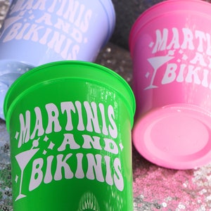 Op de afbeelding: Drie plastic bekers in pastelkleuren: lichtblauw, groen en roze. Elke beker heeft de witte tekst "MARTINIS AND BIKINIS", samen met een martini-glasafbeelding. De bekers staan op een glinsterend, met pailletten bedekt oppervlak.