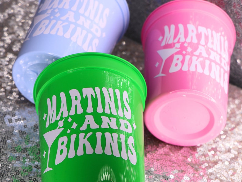 Op de afbeelding: Drie plastic bekers in pastelkleuren: lichtblauw, groen en roze. Elke beker heeft de witte tekst "MARTINIS AND BIKINIS", samen met een martini-glasafbeelding. De bekers staan op een glinsterend, met pailletten bedekt oppervlak.