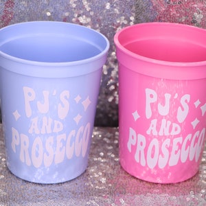 May include: Two plastic cups, one light purple and one pink, with the text "PJ's AND PROSECCO" in white. The cups are cylindrical with a slightly flared rim. The background is a sparkly, silver surface.