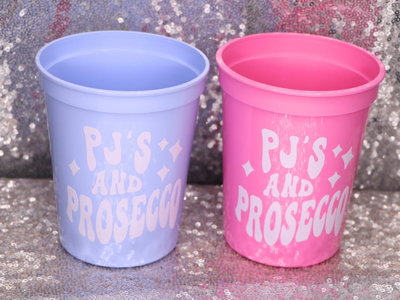 May include: Two plastic cups, one light purple and one pink, with the text "PJ's AND PROSECCO" in white. The cups are cylindrical with a slightly flared rim. The background is a sparkly, silver surface.