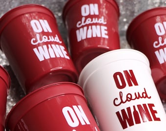 May include: A collection of red plastic cups with the phrase "ON cloud WINE" printed in white. One white cup with the same text is also visible. The cups are arranged on a reflective surface.