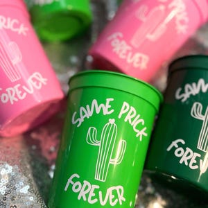May include: Several plastic cups in shades of pink and green are displayed, each with the phrase "SAME PRICK FOREVER" and a cactus illustration in white. The cups are arranged on a shiny, silver surface.