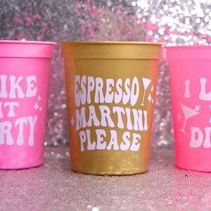 May include: Three plastic cups in pink and gold. The pink cups have the text "I LIKE IT DIRTY" in white, with a martini glass graphic. The gold cup says "ESPRESSO MARTINI PLEASE" in white. All cups have a glittery background.