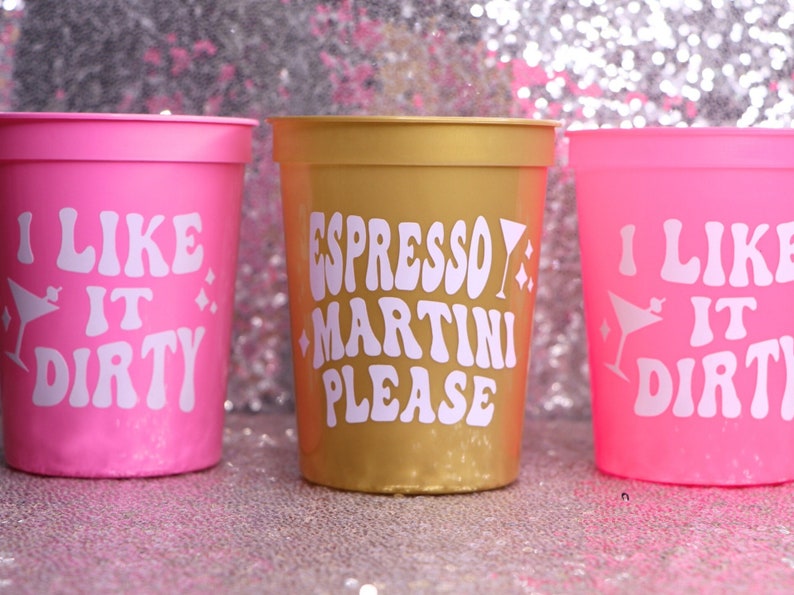 May include: Three plastic cups in pink and gold. The pink cups have the text "I LIKE IT DIRTY" in white, with a martini glass graphic. The gold cup says "ESPRESSO MARTINI PLEASE" in white. All cups have a glittery background.