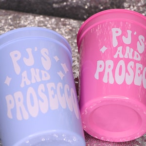 May include: Two plastic cups, one light blue and one pink, are displayed. Each cup has the words "PJ'S AND PROSECCO" in white, stylized text. The cups are on a sparkly, silver surface.