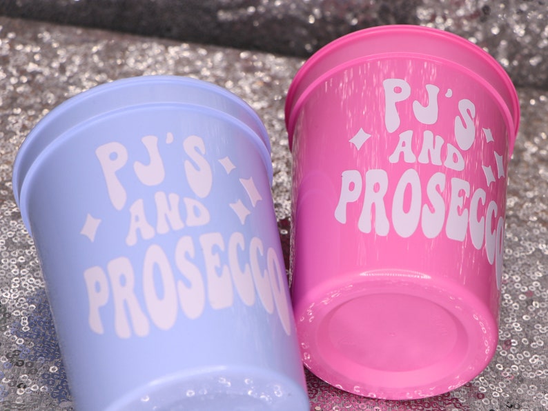 May include: Two plastic cups, one light blue and one pink, are displayed. Each cup has the words "PJ'S AND PROSECCO" in white, stylized text. The cups are on a sparkly, silver surface.