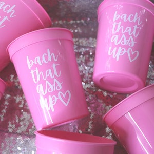 May include: Pink plastic cups with the phrase "bach that ass up" in white script, along with a heart symbol. The cups are arranged on a sparkly, pink surface, suggesting a party or celebration theme.