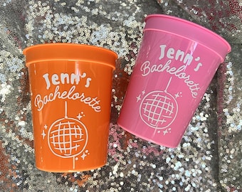 Puede incluir: Dos vasos de plástico, uno naranja y otro rosa, con el texto "Jenn's Bachelorette" y un gráfico de bola de discoteca blanco. Los vasos están sobre una superficie plateada brillante.