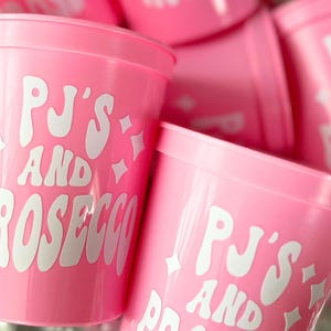 May include: Pink plastic cups with the text "PJ'S AND PROSECCO" in white, stylized lettering. The cups have a simple, cylindrical shape and are decorated with starburst graphics. The cups are stacked together.
