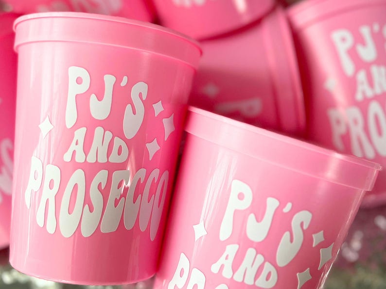 May include: Pink plastic cups with the text "PJ'S AND PROSECCO" in white, stylized lettering. The cups have a simple, cylindrical shape and are decorated with starburst graphics. The cups are stacked together.