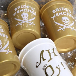May include: A collection of gold-colored plastic cups with white text and graphics. The cups feature the words "Bride's Wenches" with a skull and crossbones design. One white cup has the text "AH Oy!" with a ring graphic.