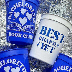 May include: Blue and white plastic cups with text for a bachelorette party. The blue cups say "Bachelorette Book Club" with hearts and an open book graphic. A white cup says "Best Chapter Yet" in blue.