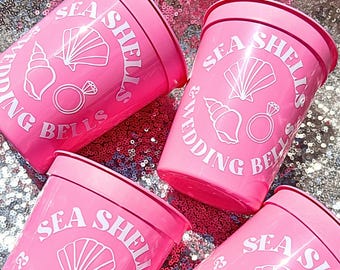 May include: Four bright pink plastic stadium cups featuring white printed graphics of a seashell, a conch shell, and a diamond ring. The text "SEA SHELLS & WEDDING BELLS" is printed in a circular arc around the central shell and ring illustrations.
