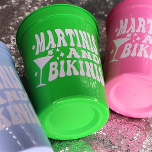 Op de afbeelding: Drie plastic bekers in lichtblauw, groen en roze. Elke beker heeft de witte tekst "MARTINIS AND BIKINIS" met een martini glas afbeelding. De bekers staan op een sprankelend, met pailletten bedekt oppervlak.