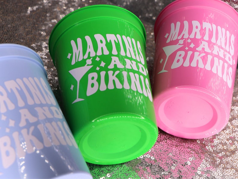 Op de afbeelding: Drie plastic bekers in lichtblauw, groen en roze. Elke beker heeft de witte tekst "MARTINIS AND BIKINIS" met een martini glas afbeelding. De bekers staan op een sprankelend, met pailletten bedekt oppervlak.