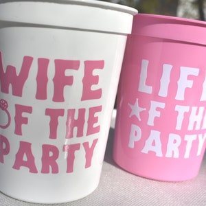 May include: Two plastic cups with pink and white text. The white cup says "Wife of the Party" with a ring icon. The pink cup says "Life of the Party" with a star icon.