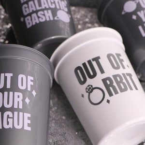 May include: Two gray plastic cups with white text that reads "Out of your league" and a small planet symbol. A white plastic cup with black text that reads "Out of orbit" and a ring symbol.
