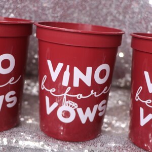 May include: Three red plastic cups with white text that reads "Vino before Vows". The cups are cylindrical with a slightly flared rim. The background is a sparkly, silver surface.