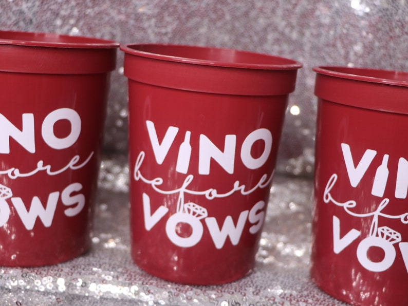 May include: Three red plastic cups with white text that reads "Vino before Vows". The cups are cylindrical with a slightly flared rim. The background is a sparkly, silver surface.