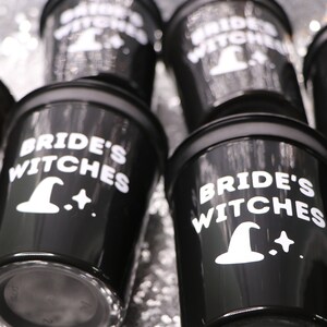 May include: Black shot glasses with the words "Bride's Witches" in white, along with a witch hat and star graphic. The glasses have a glossy finish and are arranged on a shiny, silver surface. Perfect for a bachelorette party or Halloween celebration.
