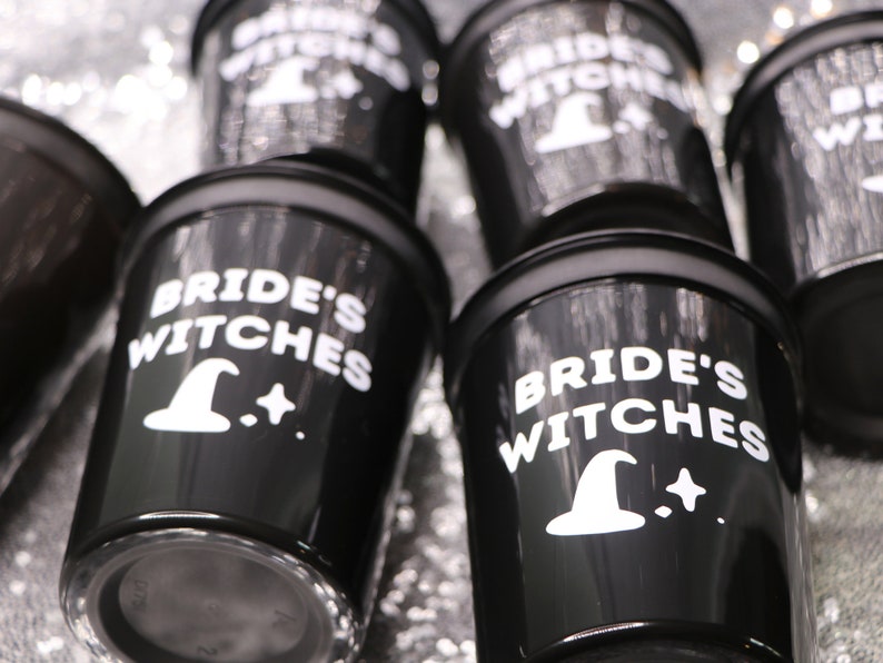 May include: Black shot glasses with the words "Bride's Witches" in white, along with a witch hat and star graphic. The glasses have a glossy finish and are arranged on a shiny, silver surface. Perfect for a bachelorette party or Halloween celebration.