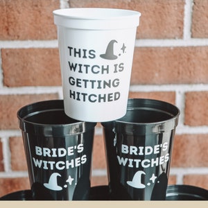 May include: White and black plastic cups with witch-themed text and graphics. One cup reads "THIS WITCH IS GETTING HITCHED" with a witch hat and star. Other cups say "BRIDE'S WITCHES" with a witch hat and star. The cups are stacked.