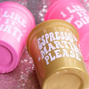 May include: Three plastic cups with white text. Two are bright pink and say "I LIKE IT DIRTY". The third is gold and says "ESPRESSO MARTINI PLEASE". The cups are on a sparkly pink surface.