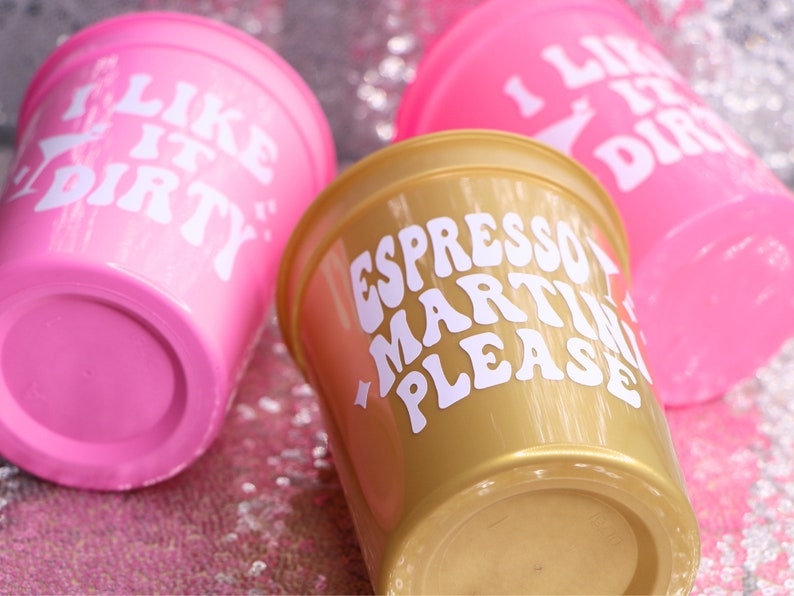 May include: Three plastic cups with white text. Two are bright pink and say "I LIKE IT DIRTY". The third is gold and says "ESPRESSO MARTINI PLEASE". The cups are on a sparkly pink surface.