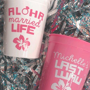 May include: Two pink and white plastic cups with a hibiscus flower design. One cup says "Aloha married life" and the other says "Michelle's Last Luau".