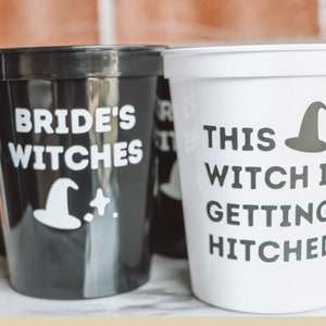 May include: Black and white plastic cups with text and witch hat graphics. One cup reads "Bride's Witches" and another says "This Witch Is Getting Hitched". The cups are likely for a bachelorette party or bridal shower.
